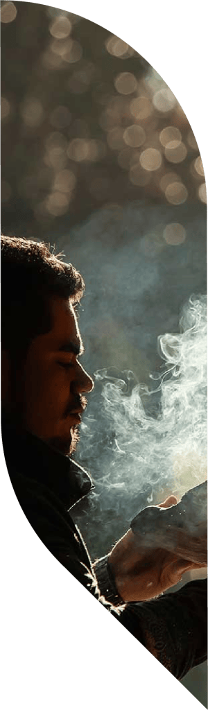 Aboriginal man blowing on leaves with smoke rising